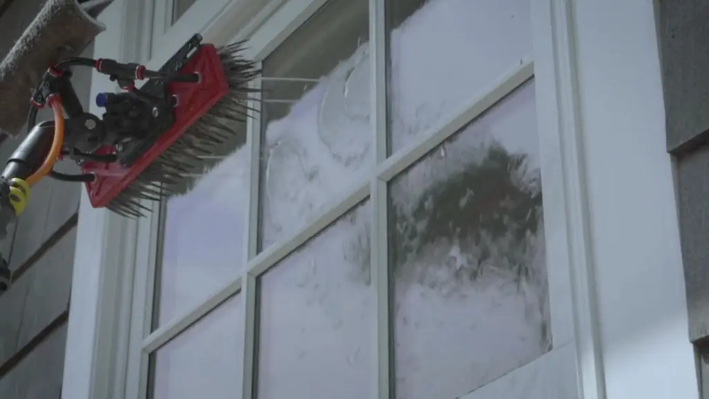 A water-fed brush scrubbing divided-light panes set in white trim beside cedar shingles, half the glass still clouded