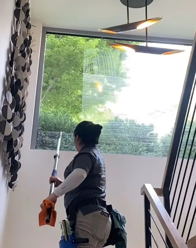 A tech cleaning a tall interior picture window above a stair landing